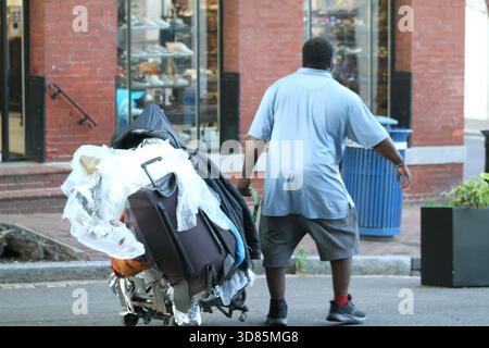 Homme portant un chariot rempli de bagages lourds et d'autres effets personnels sur une route de ville dans les États-Unis Banque D'Images