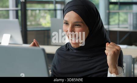 Islamique ethnique heureuse femme d'affaires gagnant en ligne célébration de réalisation femme d'affaires dans le hijab dans le bureau gagner victoire ordinateur Internet bonne nouvelle Banque D'Images