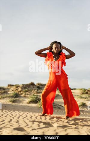Jeune femme noire avec des tresses posant pieds nus sur les dunes de sable du désert dans une combinaison plissée orange vibrante, bijoux élégants et plein air confiant et élégant Banque D'Images