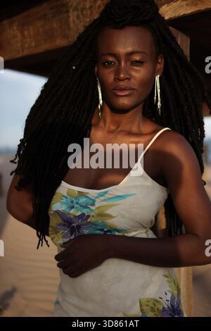 Jeune femme noire avec des dreadlocks naturels et un regard confiant se penche sous une structure de plage en bois à l'heure dorée, portant une robe florale et la lumière du soleil Banque D'Images