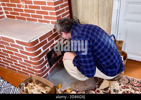Empilant le bois de chauffage dans le foyer de poêle en brique, le propriétaire prépare le chauffage à bois avec banc à l'intérieur de la maison de campagne avant d'allumer les bûches. Banque D'Images
