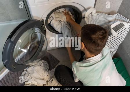 Garçon avec des lunettes chargeant des vêtements sales dans la machine à laver à partir du panier à linge dans la salle de bains à la maison vue de dessus. Concept des enfants faisant du chor de ménage Banque D'Images