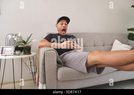 Homme réagissant avec une expression choquée en regardant un téléphone portable sur un canapé de salon Banque D'Images