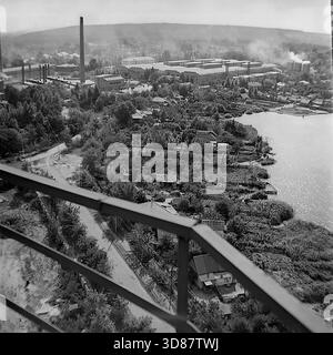 Slavyansk, Ukraine, URSS - années 1970 : vue en grand angle de l'usine d'armature-isolant AIZ et de la zone résidentielle adjacente. Les poutres métalliques floues encadrent le premier plan. Ci-dessous, des maisons à un étage avec jardins bordent la rive. En arrière-plan, le complexe industriel massif avec une grande cheminée en briques et des évents de fumée domine le paysage. Cette photo d'archives en noir et blanc capture la coexistence de l'industrie lourde et de la vie rurale privée dans la paisible ville industrielle de Donbass. Banque D'Images