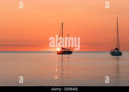 Deux voiliers amarrés sur une mer calme ressemblant à du verre, silhouettés contre le ciel orange intense au lever ou au coucher du soleil sur l'horizon Banque D'Images