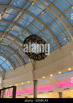 Une grande couronne de Noël illuminée est suspendue à un plafond de lucarne en verre à l'intérieur d'un bâtiment moderne, ornée de lumières festives et de détails architecturaux Banque D'Images