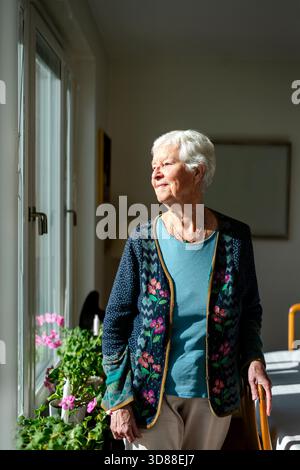 Femme âgée souriante regardant par la fenêtre tout en se tenant debout dans le salon à la maison Banque D'Images