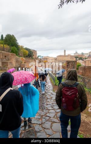 Un groupe de touristes marchent le long d'un chemin pavé sous la pluie, en utilisant des parapluies et des vêtements de pluie pour rester au sec tout en explorant les ruines antiques de R. Banque D'Images