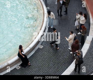 Une femme en robe noire est assise au bord de la fontaine de Trevi tandis qu’un photographe la prend en photo. D'autres personnes se tiennent debout à regarder Banque D'Images