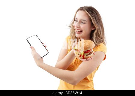 Joyeuse jeune femme souriante profitant de la livraison de nourriture, tenant un hamburger dans une main et un smartphone dans l'autre, isolée sur un fond blanc. Onlin Banque D'Images