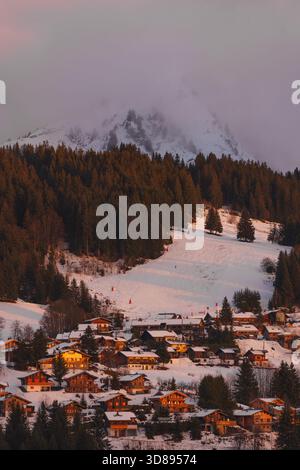 Les Gets, haute-Savoie, France – 26 décembre 2022 : la lumière du coucher du soleil réchauffe les chalets alpins sous une piste de ski couverte de brume dans les Alpes françaises Banque D'Images