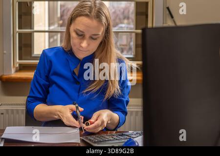 Femme en robe bleue est engagée dans la coupe de papier à un bureau, avec des fournitures de bureau et une calculatrice à proximité, illustrant un travail créatif et productif Banque D'Images