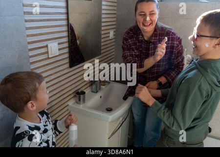 Heureuse mère et deux fils avec de la crème sur les visages riant ensemble dans la salle de bain pendant la routine de soins de la peau du matin près de l'évier. Concept de plaisir en famille, parentin Banque D'Images