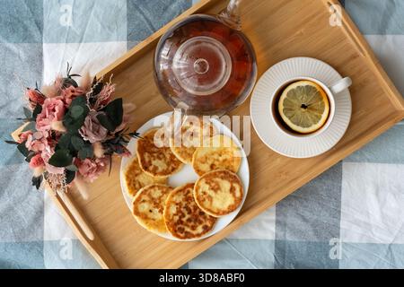 Plateau en bois avec crêpes au fromage cottage sur assiette, tasse à thé avec citron, théière en verre et fleurs sur nappe à carreaux vue de dessus. Concept de brea confortable Banque D'Images