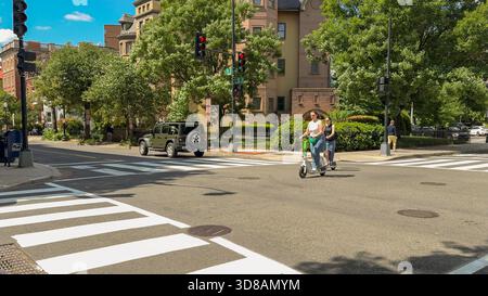 Washington D.C. États-Unis - 24 mai 2025. Deux femmes sur scooters électriques traversent une rue ensoleillée entourée d'architecture historique et de verdure. Banque D'Images