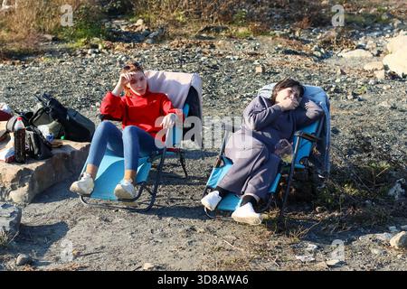 Plan large de jeune femme en rouge et femme plus âgée en gris assis détendu dans des chaises sur un sol pierreux, avec des équipements de plein air à proximité. Banque D'Images