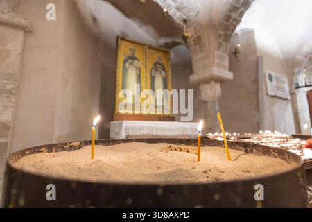 De fines bougies de cire d'abeille brûlent dans le sable dans la crypte de l'établissement Nicholas, Bari, sur fond flou d'arches romanes et d'une icône dorée. Banque D'Images
