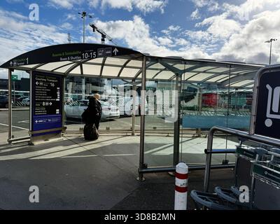 Dublin, Irlande - 21 octobre 2025 : un voyageur avec une valise roulante attend à l'abri en verre de l'aéroport de Dublin à côté d'une zone d'arrivée très fréquentée. Banque D'Images