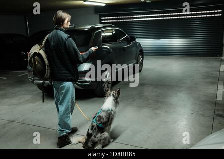 Une femme se tient debout dans un garage souterrain avec son chien, tenant une clé de voiture près d'un véhicule stationné. La scène montre la préparation pour la conduite, le voyage des animaux de compagnie Banque D'Images