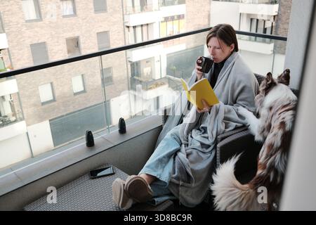 Une femme enveloppée dans une couverture lit un cahier sur un balcon tout en sirotant une boisson chaude, tandis que son chien se tient à proximité. Le moment reflète le calme, le confort Banque D'Images