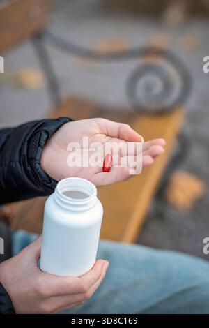 Une capsule rouge sur une paume au-dessus d'une bouteille blanche ouverte dans un cadre extérieur calme. Maquette, mise au point sélective. Banque D'Images