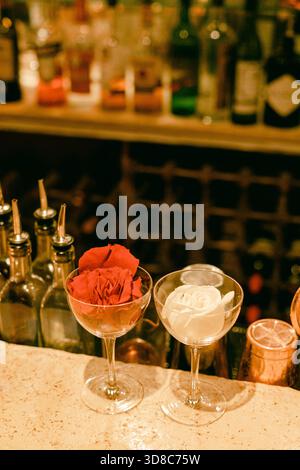 Deux cocktails élégants avec des roses sont assis sur un bar, entouré de diverses bouteilles de liqueur. Banque D'Images