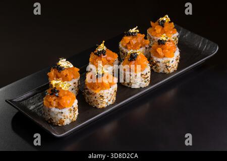 Élégants rouleaux de sushi recouverts de saumon épicé, de caviar noir et de luxueux flocons de feuilles d'or sur une assiette noire, un plat gastronomique parfait pour une cuisine raffinée. Banque D'Images