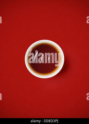 Vue plate de haut en bas de la tasse en céramique blanche remplie de thé oolong foncé isolé sur fond rouge texturé Banque D'Images
