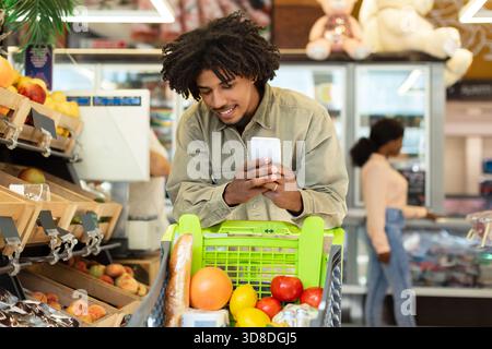 Jeune homme vérifie le téléphone tout en magasinant pour l'épicerie dans un supermarché dynamique Banque D'Images