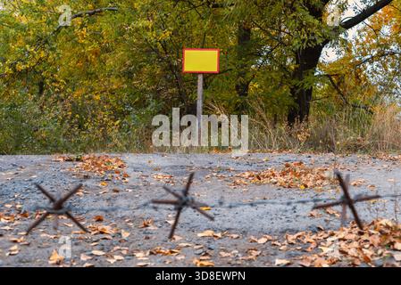 Une clôture de barbelés s'étend sur un chemin de gravier, menant à un panneau blanc entouré d'un feuillage d'automne coloré. Banque D'Images