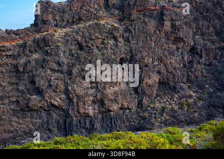 Face de falaise volcanique massive avec des formations de basalte en couches et des strates minérales rouges dans le paysage d'Anaga Banque D'Images