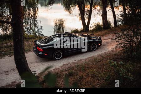 Noir Pontiac Firebird sur une route rurale au coucher du soleil. Vue latérale aérienne d'une voiture de sport américaine sur un chemin de terre près d'un lac dans les rayons du soleil couchant. Banque D'Images