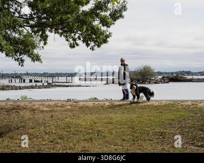 Une femme se tient près de la rive rocheuse de la rivière Delaware dans le New Castle Battery Park avec son chien bernedoodle noir et blanc. Banque D'Images