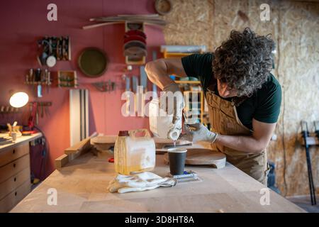 Menuisier homme préparant pour le travail sur le nouvel objet de mobilier, versant la quantité de résine époxy dans la tasse de papier Banque D'Images