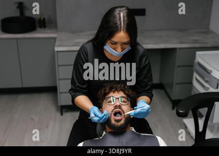 Dentiste femme portant un masque et des gants effectuant un bilan dentaire sur un patient masculin dans une clinique moderne. Se concentrer sur l'hygiène bucco-dentaire Banque D'Images