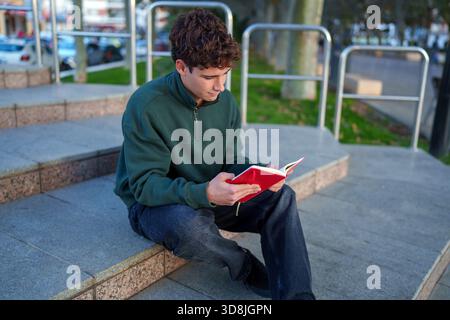 Étudiant aux cheveux bouclés est assis sur des marches de granit à l'extérieur, immergé dans un livre rouge, embrassant pleinement l'atmosphère éducative sur le campus Banque D'Images