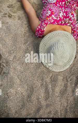 Femme asiatique en robe de plage colorée couchée en faisant une sieste sur le sable de plage couvrant son visage avec chapeau crochet rond pendant la retraite d'été et les vacances Banque D'Images