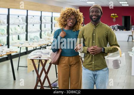 Jeune adulte femme noire et jeune adulte homme noir debout ensemble dans le magasin, souriant et regardant la caméra, tenant le panier et reliant les bras, étagères avec la vaisselle en arrière-plan Banque D'Images