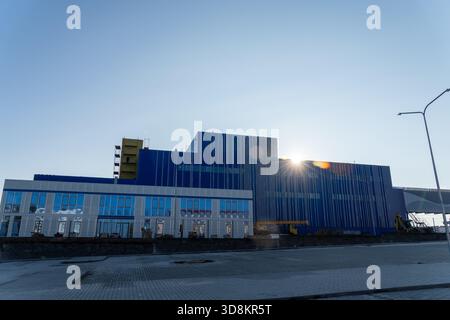 Un grand bâtiment industriel moderne avec une façade bleue vibrante se dresse sous un ciel clair, avec le soleil éclatant derrière la structure pendant const Banque D'Images