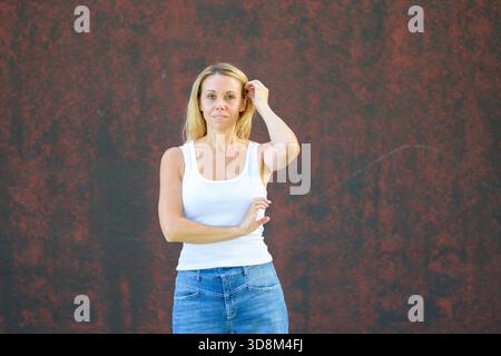 Femme blonde en débardeur blanc décontracté et jeans debout à l'extérieur contre un mur rouge texturé, posant naturellement avec une confiance calme. Banque D'Images