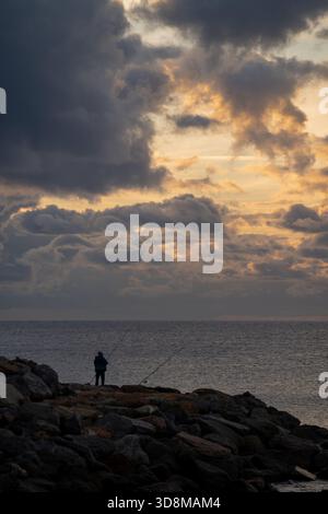 silhouette près du rivage du lever du soleil, individu debout tranquillement sur des jetées accidentées à l'aube, personne seule s'engage dans un rituel de pêche matinale paisible à Banque D'Images