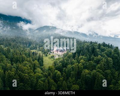 Vue aérienne du majestueux château de Peles niché au milieu de forêts verdoyantes, avec des montagnes spectaculaires entourées de brume s'élevant en arrière-plan, Sinaia, RO Banque D'Images