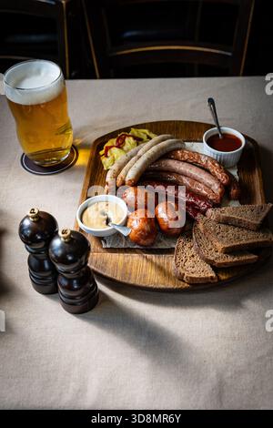 Plan vertical d'un plateau en bois avec des saucisses grillées, des trempettes, du pain et un verre de bière sur une table. Banque D'Images