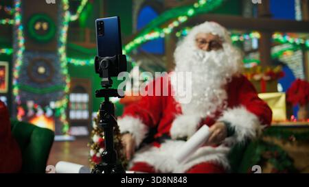 Santa Claus utilisant un smartphone sur trépied pour filmer les lettres d'ouverture de vlog des enfants. Personne âgée portant le costume de Noël enregistrant la vidéo avec la caméra selfie du téléphone, lisant les lettres des enfants, caméra A. Banque D'Images