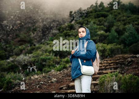 Randonneur femme en veste bleue et sac à dos se tient en toute confiance sur un sentier pittoresque, entouré d'une verdure vibrante et d'un terrain rocheux, incarnant adv Banque D'Images