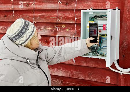 Capture des relevés de compteurs résidents ruraux dans des vêtements chauds utilise le téléphone photographiant le compteur d'énergie à l'intérieur du panneau résidentiel sur le mur extérieur de la maison. Banque D'Images