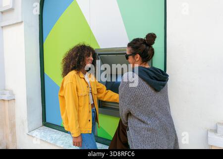 Deux femmes multiraciales heureuses utilisant un guichet automatique pour une transaction financière, partageant un moment et une discussion en plein air Banque D'Images