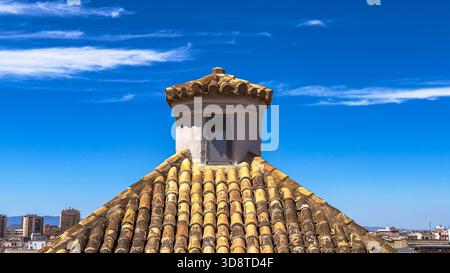 Toit méditerranéen traditionnel avec tuiles d'argile rustique et petite tour de cheminée blanche contre le ciel bleu vif et les nuages doux par jour ensoleillé. Banque D'Images