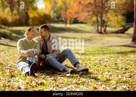 Une femme coréenne et un homme caucasien sont assis ensemble sur l'herbe, riant et profitant d'une journée d'automne ensoleillée entourée de feuilles colorées. Banque D'Images