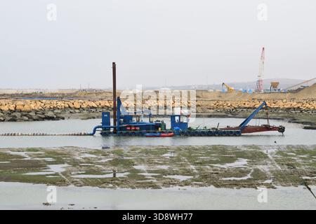 l'image montre un navire de dragage bleu ou une drague aspirante de coupe travaillant dans une zone côtière ou portuaire peu profonde, avec une longue échelle ou un tuyau s'étendant vers l'avant Banque D'Images